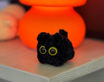 May include: A black crocheted plush toy cat with yellow eyes. The cat is sitting on a white shelf in front of an orange lamp.