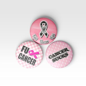 May include: Three pink button pins with different designs. The first pin has a pink ribbon with a floral design and the text "Fight Warrior Fight". The second pin has a pink ribbon and the text "FU CANCER" on a pink and white checkered background. The third pin has a pink ribbon pattern and the text "CANCER SUCKS".
