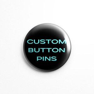 May include: A circular button pin with a glossy black finish. The text CUSTOM BUTTON PINS is printed in the center in a bold, light blue sans-serif font. The pin has a smooth, reflective surface and a clean, rounded edge design.