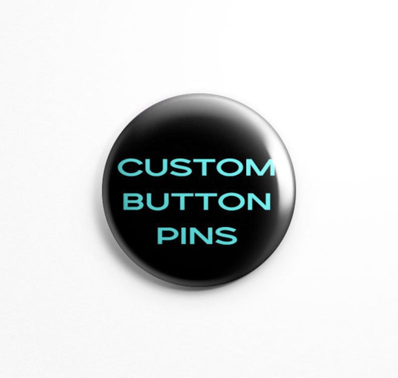 May include: A circular button pin with a glossy black finish. The text CUSTOM BUTTON PINS is printed in the center in a bold, light blue sans-serif font. The pin has a smooth, reflective surface and a clean, rounded edge design.