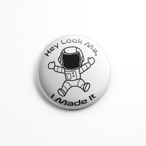 May include: White button with a black and white illustration of an astronaut with the text "Hey Look Ma, I Made It"
