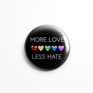 May include: A black button with the text "MORE LOVE LESS HATE" in white. There are six rainbow-colored hearts below the text.
