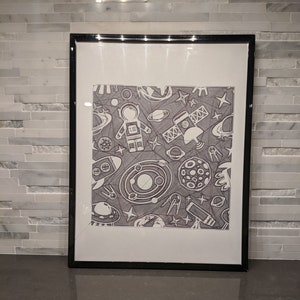 May include: A black and white framed print of a space-themed illustration. The print features an astronaut, planets, stars, and a satellite.