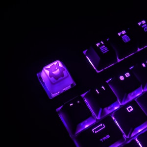 以下が含まれることがあります： 紫色のバックライト付きキーボードで、1つのキーキャップに漫画のモンスターのデザインが施されています。キーキャップは明るい紫色の光で照らされています。