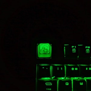 May include: A green illuminated keyboard with a dinosaur icon on the top left key. The keys are labeled with numbers and letters, including F1, F2, F3, 1, 2, 3, 4, Q, W, E, and TAB.