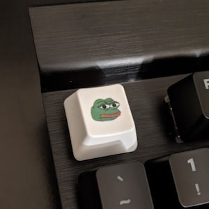 May include: A black keyboard with a white keycap featuring a green Pepe the Frog meme. The keycap is labeled with the letter "F".