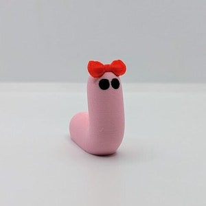 May include: A pink worm with black eyes and a red bow on its head. The worm is made of clay and is a handmade toy.