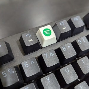 May include: A black keyboard with a white keycap featuring the Spotify logo. The keycap is located above the F5, F7, and F8 keys. The keyboard has black keys with white lettering for the numbers, symbols, and letters.