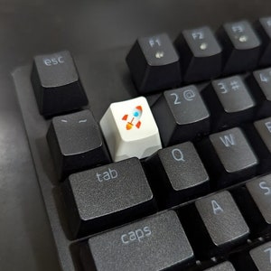 Peut inclure: Un clavier noir avec une touche blanche représentant une fusée. La touche est située sur la rangée supérieure du clavier, entre les touches "esc" et "tab".
