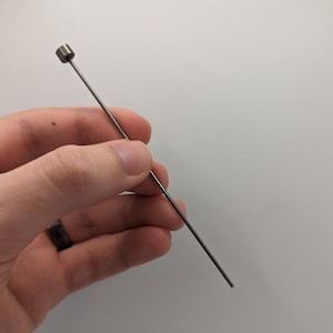 May include: A thin, silver-colored metal rod with a rounded tip. The rod is held in a hand.