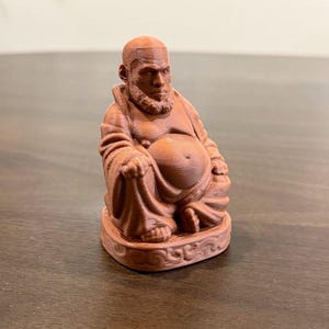 May include: A brown figurine of a seated man with a beard and bald head. The figure is wearing a robe and has a large belly. The base of the figurine has a decorative, swirling pattern.