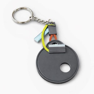 Peut inclure: Un porte-clés moderne avec un grand pendentif circulaire gris foncé. Le porte-clés comprend un porte-clés et une chaîne argentés, avec des accents colorés de bleu, jaune et orange. Le pendentif a un trou et un petit détail argenté.