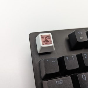 May include: A white keyboard keycap with a printed image of a man's face with a moustache and a surprised expression.
