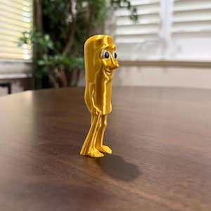 May include: A 3D-printed, golden-yellow figurine of a cartoon character with a rectangular body, thin legs, and small feet. The character has a smiling face with blue eyes and a prominent nose. The figurine is standing on a dark brown surface.