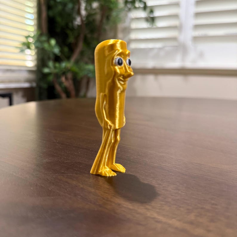 May include: A 3D-printed, golden-yellow figurine of a cartoon character with a rectangular body, thin legs, and small feet. The character has a smiling face with blue eyes and a prominent nose. The figurine is standing on a dark brown surface.