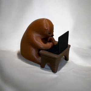 May include: A brown bear-like figurine is seated at a small brown table, typing on a black laptop. The bear has a rounded body and is focused on the computer. The figurine is made of a solid material and has a smooth texture. The background is a soft white.
