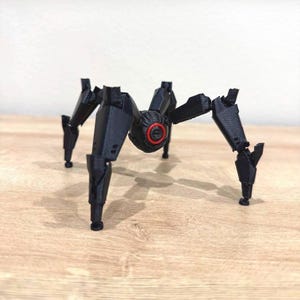 May include: A black, mechanical spider model with articulated legs and a central body. The body features a red circular detail. The model is displayed on a wooden surface.