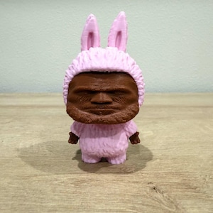 Puede incluir: Figura impresa en 3D de una persona con un disfraz de conejito rosa. La figura tiene una cara y manos marrones, con una capucha y orejas de conejo rosas y esponjosas. La figura está sobre una superficie de madera.