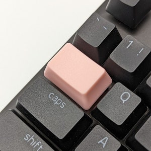 Op de afbeelding: Een zwart toetsenbord met een roze toetsdop met het label "caps". Het toetsenbord heeft toetsen met de labels "esc", "~", "1!", "Q", "A" en "shift".