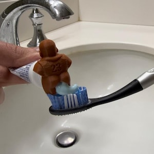 Può includere: Uno spazzolino da denti originale con manico nero e setole blu, che eroga dentifricio. Una statuetta marrone con il numero 23 è attaccata al tubetto di dentifricio. La scena è ambientata in un bagno con un lavandino bianco e un rubinetto cromato.
