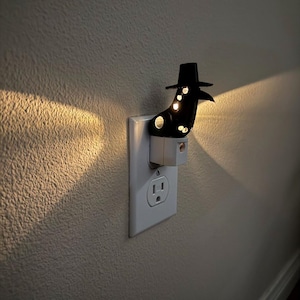 Op de afbeelding: Een zwart nachtlampje in de vorm van een pestdoktermasker en hoed, aangesloten op een wit stopcontact. Het licht schijnt door de oog- en snavelopeningen van het masker en werpt een warme gloed op de muur. Een decoratief en functioneel item.