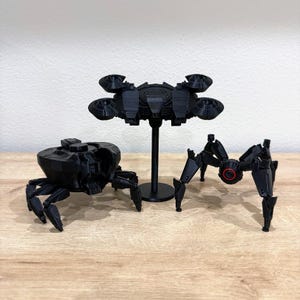 May include: Three black 3D-printed models of futuristic robots. One is a spider-like robot with articulated legs, another is a drone-like model on a stand, and the third is a crab-like robot with multiple legs.