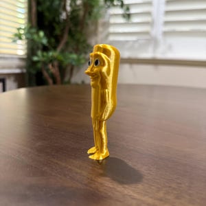 May include: A 3D-printed, golden-yellow figurine of a cartoonish character with a long nose, small eyes, and a smiling expression. The figure is standing upright with legs and feet visible. The background is blurred, with a wooden table and a window.