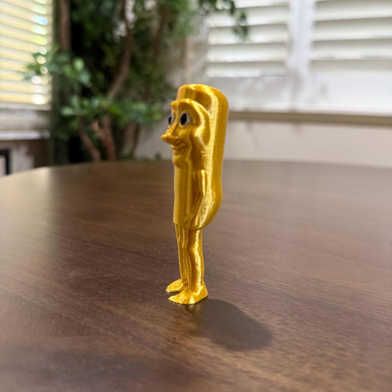 May include: A 3D-printed, golden-yellow figurine of a cartoonish character with a long nose, small eyes, and a smiling expression. The figure is standing upright with legs and feet visible. The background is blurred, with a wooden table and a window.