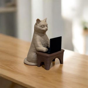 May include: A beige cat figurine seated at a small brown table, appearing to work on a black laptop. The cat is in a seated position, with its paws on the keyboard. The figurine is on a wooden surface.
