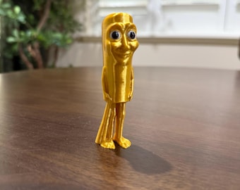 May include: A golden-colored, 3D-printed figurine of a smiling, anthropomorphic object with a cylindrical body, small feet, and googly eyes. The figure stands on a dark brown wooden surface, with a blurred green plant in the background.