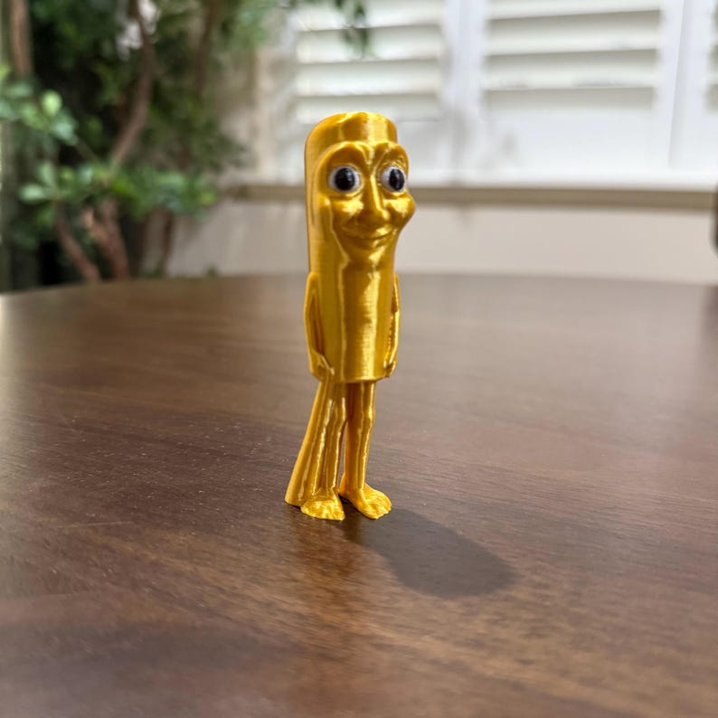 May include: A golden-colored, 3D-printed figurine of a smiling, anthropomorphic object with a cylindrical body, small feet, and googly eyes. The figure stands on a dark brown wooden surface, with a blurred green plant in the background.
