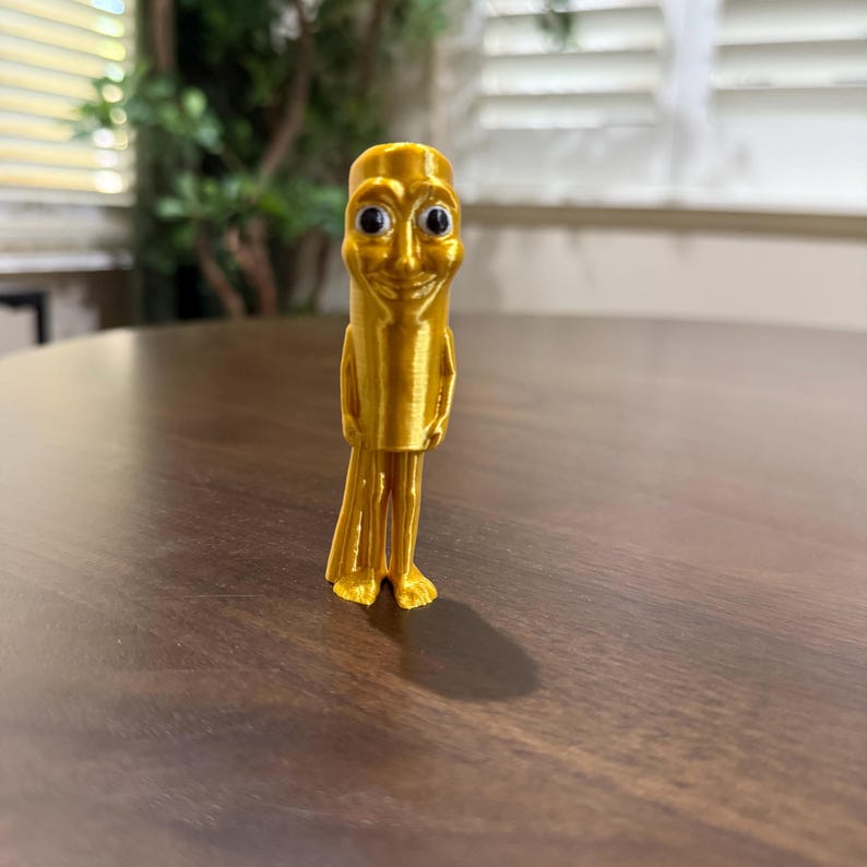 May include: A gold-colored, 3D-printed figurine of a smiling humanoid figure. The figure has a cylindrical body, small arms, and a skirt-like lower portion. The eyes are black, and the figure is standing on a wooden surface.