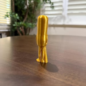 May include: A gold-colored, 3D-printed figurine of a stylized figure with a rounded head and elongated body. The figure has thin, leg-like structures at the base. The figurine is positioned on a dark brown wooden surface.