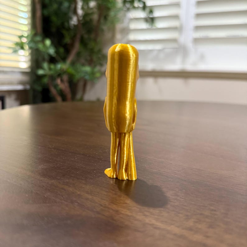 May include: A gold-colored, 3D-printed figurine of a stylized figure with a rounded head and elongated body. The figure has thin, leg-like structures at the base. The figurine is positioned on a dark brown wooden surface.