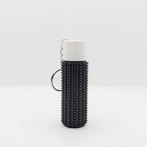 Op de afbeelding: Een zwart-witte sleutelhanger met een getextureerd, stekelig ontwerp. De sleutelhanger heeft een zilveren ring en een witte bovenkant.