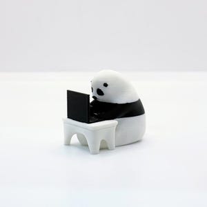 Puede incluir: Una figurita de panda blanca y negra sentada en una mesa blanca pequeña, usando una computadora portátil negra.