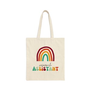 May include: A white canvas tote bag with a rainbow design and the text "research assistant" in colorful, retro-style lettering.
