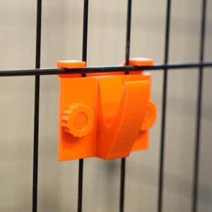 May include: Bright orange plastic hook attached to a black wire fence. The hook has a V-shape and two circular knobs. The hook is designed to attach to a wire fence and hold an item. The background is a blurred neutral color.