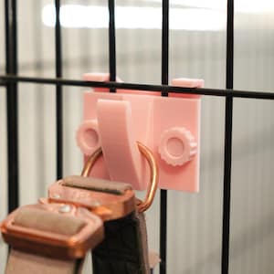 Peut inclure: Un crochet en plastique rose à clipser avec un anneau circulaire, fixé à une grille métallique noire. Un collier de chien avec une boucle de couleur cuivre est suspendu au crochet. Le crochet a deux boutons circulaires pour le réglage.