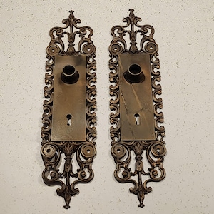 May include: Two ornate brass door plates with intricate scrollwork and a keyhole. The plates are rectangular and have a raised design.