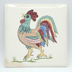 May include: A square ceramic tile featuring a hand-painted rooster illustration. The rooster has a light blue head, a red comb and wattle, and a tan body. Its tail feathers are multicolored in shades of blue, yellow, red, and purple.