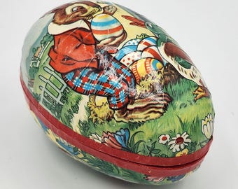 May include: Vintage Easter egg-shaped container with a red rim. The egg features a colorful illustration of a rabbit, duck, and decorated Easter eggs on a green grassy background with flowers. The egg is made of a paper-like material.