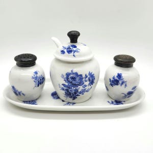 May include: A white porcelain condiment set with blue floral designs. The set includes a lidded sugar bowl with a spoon, and two shakers with black lids, all on a matching oval tray. The floral pattern is repeated on each piece.