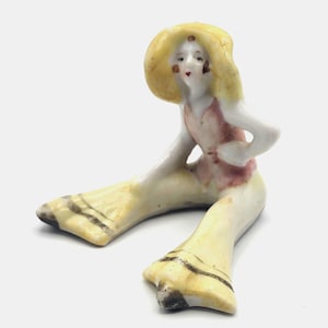 May include: A vintage porcelain figurine of a seated woman wearing a yellow hat and pants, and a pink top. The figurine has a hand-painted face with red lips and blush. The pants have dark brown stripes near the bottom.