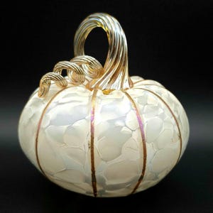 May include: A decorative white glass pumpkin with gold accents. The pumpkin has a textured surface and gold vertical lines. The stem is a twisted gold color. The pumpkin is a fall-themed decorative item.