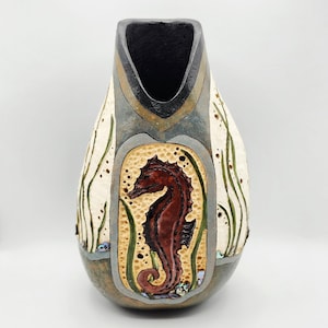 May include: A teardrop-shaped gourd vessel featuring a dark brown seahorse relief centered on a textured tan background. The exterior is decorated with painted green seaweed, blue accents, and dark gray borders, with a black interior opening.