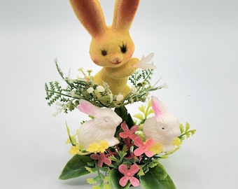 May include: A decorative topper featuring a yellow flocked rabbit positioned above two smaller white rabbits. The rabbits are surrounded by green foliage, small white flowers, and pink blossoms, all mounted on a clear plastic stake for display.