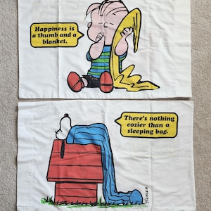 May include: Two white pillowcases featuring Peanuts characters. One shows Linus with his thumb and blanket, the text bubble reads "Happiness is a thumb and a blanket." The other shows Snoopy on his doghouse, with the text "There's nothing cozier than a sleeping bag."