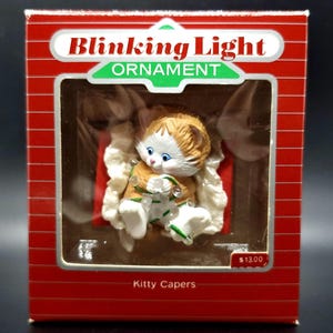 May include: A white and orange kitten wearing a green and white outfit with a string of lights is sitting in a white rocking chair. The ornament is in a red box with the words "Blinking Light Ornament" and "Kitty Capers" on it. The price is $13.00.