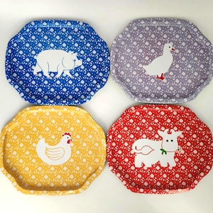 May include: A set of four scalloped-edge rectangular trays featuring a white floral pattern on solid backgrounds. The blue tray displays a white pig, the lavender tray a white goose, the yellow tray a white chicken, and the red tray a white cow.